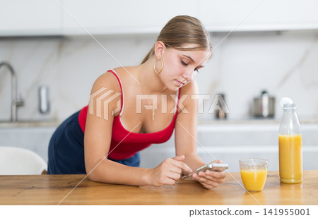 Teenage girl looking at smartphone while drinking fresh juice in kitchen Teenage girl looking at smartphone while drinking fresh juice in kitchen 141955001