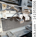 Detailed close-up of a disassembled car front end showing the radiator and A/C condenser. Also visible are a dusty horn, wiring harnesses, metal frame, and plastic brackets 141955006