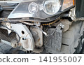 Close-up view of a disassembled car front end. Exposes a dusty headlight assembly, metal frame structure, radiator, transmission cooler, and complex wiring harnesses. Likely awaiting repair. 141955007