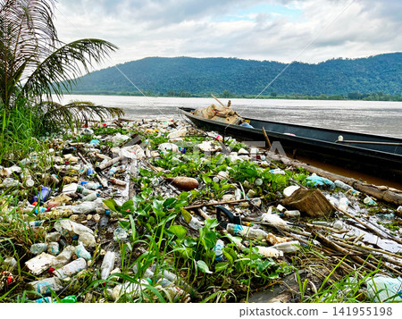 Massive plastic garbage and water hyacinth weed accumulate along the natural riverbank near wooden boat, highlighting severe environmental pollution crisis Massive plastic garbage and water hyacinth weed accumulate along the natural riverbank near wooden boat, highlighting severe environmental pollution crisis 141955198