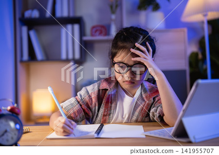 Young girl with glasses thinking hard while studying late at night with laptop and notebook in cozy room lighting and focused expression 141955707