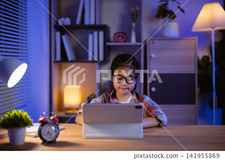 Young girl wearing glasses sitting at wooden desk using tablet device in cozy modern room with warm light during evening Young girl wearing glasses sitting at wooden desk using tablet device in cozy modern room with warm light during evening 141955869