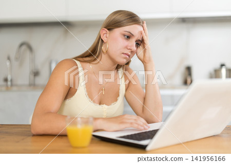 teenager is sitting at a table in the kitchen and sadly looking at a laptop screen teenager is sitting at a table in the kitchen and sadly looking at a laptop screen 141956166