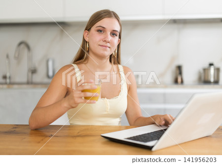 Happy teenage girl working on laptop and drinking juice in kitchen Happy teenage girl working on laptop and drinking juice in kitchen 141956203