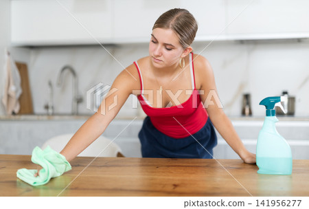 Teenage girl wiping wooden surfaces with cleaner in kitchen Teenage girl wiping wooden surfaces with cleaner in kitchen 141956277