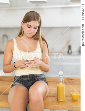 Attentive teenage girl looking at smartphone while standing in kitchen Attentive teenage girl looking at smartphone while standing in kitchen 141956321