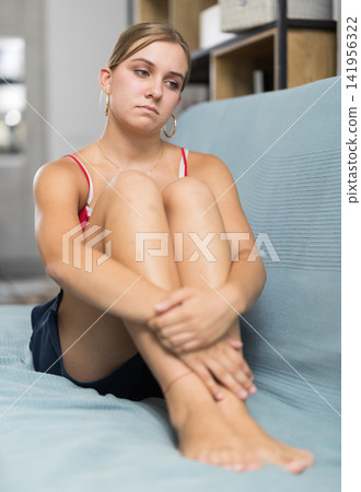 Sad girl sitting on sofa at home 141956322