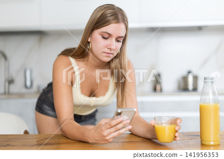 Teenage girl standing in the kitchen, drinking juice and scrolling through social networks on mobile Teenage girl standing in the kitchen, drinking juice and scrolling through social networks on mobile 141956353