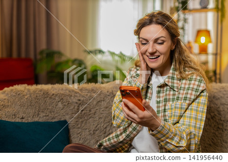 Young woman on home sofa reading delivery message on smartphone celebrating online game win with joy Young woman on home sofa reading delivery message on smartphone celebrating online game win with joy 141956440