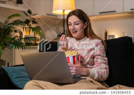 Adult woman with laptop and popcorn watches trailer movie online laughs comments comedy fun at night 141956455