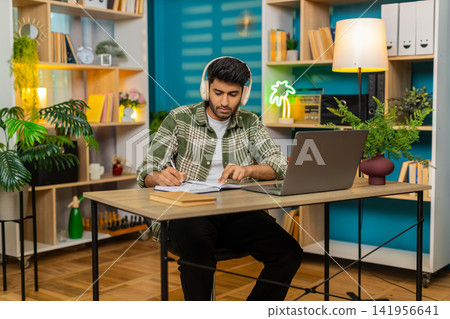 Indian man on home table taking notes in headphones during online lesson with serious focused face 141956641