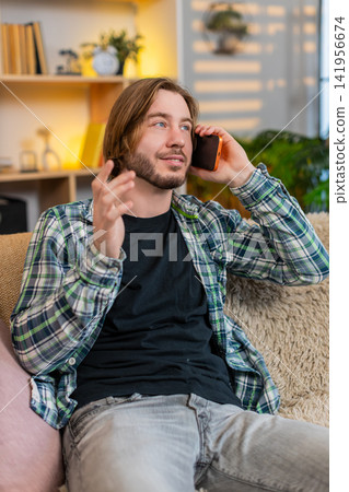 Young man on home sofa talking on smartphone and gesturing during cheerful phone call in living room Young man on home sofa talking on smartphone and gesturing during cheerful phone call in living room 141956674