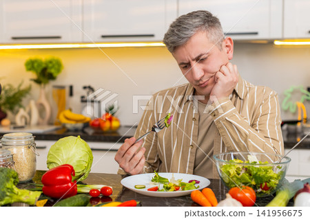 Middle-aged man eating disliked vegetable salad in kitchen during strict diet with strong willpower 141956675