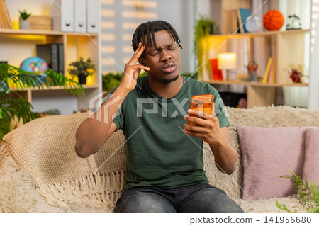 African American young man on home sofa feeling upset at bad smartphone news after online game loss 141956680