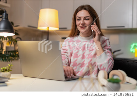Adult woman talks on smartphone while working on laptop at night voice call smiles productive chat 141956681
