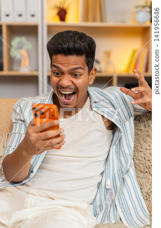 Indian man on home sofa shocked by smartphone feed in living room with happy wow surprise reaction Indian man on home sofa shocked by smartphone feed in living room with happy wow surprise reaction 141956686