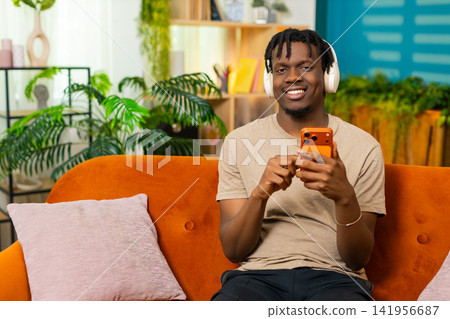 African American young man on home sofa putting on headphones and starting music on smartphone app African American young man on home sofa putting on headphones and starting music on smartphone app 141956687