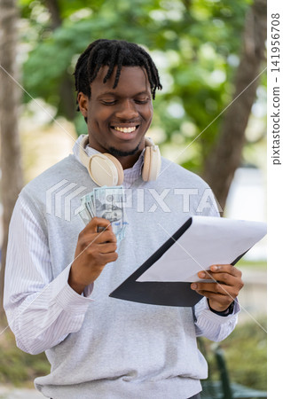 African American young man smiling with cash and documents after successful deal in city street park African American young man smiling with cash and documents after successful deal in city street park 141956708