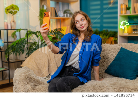 Young woman on home sofa takes selfie with smartphone checking cute pose and smiling in living room Young woman on home sofa takes selfie with smartphone checking cute pose and smiling in living room 141956734