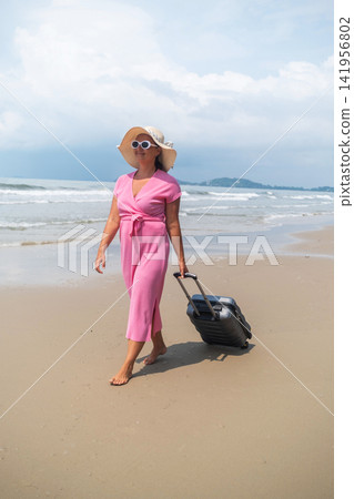 Young woman in pink dress and large straw hat walking with a rolling grey suitcase beside gentle sea waves on a sunny sandy beach. Summer travel concept Young woman in pink dress and large straw hat walking with a rolling grey suitcase beside gentle sea waves on a sunny sandy beach. Summer travel concept 141956802