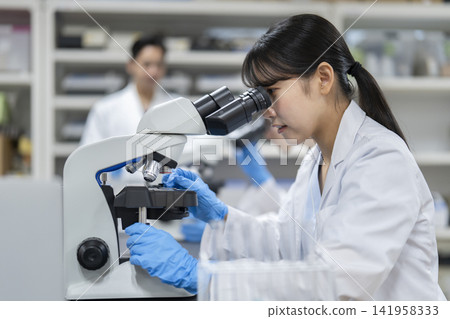 A young woman in a white coat working with a microscope (profile view) A young woman in a white coat working with a microscope (profile view) 141958333