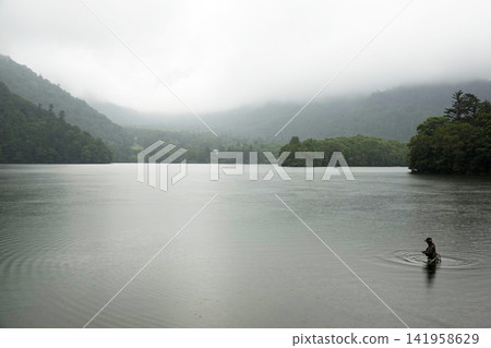 A fisherman wading in a lake with misty mountain ranges and a mystical atmosphere. A fisherman wading in a lake with misty mountain ranges and a mystical atmosphere. 141958629