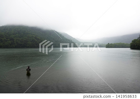 A fisherman wading in a lake with misty mountain ranges and a mystical atmosphere. A fisherman wading in a lake with misty mountain ranges and a mystical atmosphere. 141958633