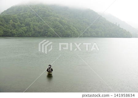 A fisherman wading in a lake with misty mountain ranges and a mystical atmosphere. A fisherman wading in a lake with misty mountain ranges and a mystical atmosphere. 141958634