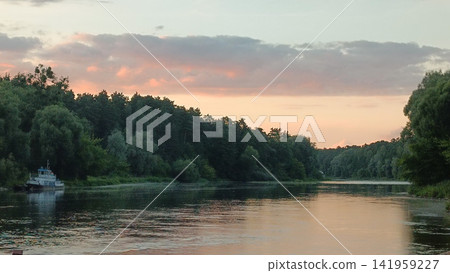 A peaceful dusk landscape with a wide calm river mirroring warm orange and pink sky colors, a moored white-and-blue boat near the bank, and dense green trees lining the water. A peaceful dusk landscape with a wide calm river mirroring warm orange and pink sky colors, a moored white-and-blue boat near the bank, and dense green trees lining the water. 141959227