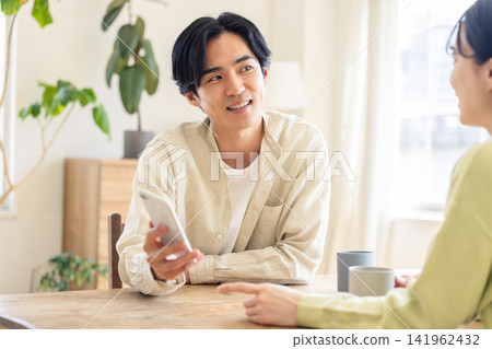 一對三十多歲的夫婦正在一起操作智慧型手機,一邊看著螢幕一邊微笑聊天。 一對三十多歲的夫婦正在一起操作智慧型手機,一邊看著螢幕一邊微笑聊天。 141962432