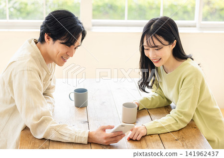 A couple in their 30s are seen operating a smartphone together, smiling and chatting while looking at the screen. A couple in their 30s are seen operating a smartphone together, smiling and chatting while looking at the screen. 141962437