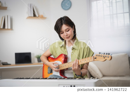 Young woman playing electric guitar in cozy living room, practicing music, smiling, with keyboard and open book nearby, relaxed indoor atmosphere Young woman playing electric guitar in cozy living room, practicing music, smiling, with keyboard and open book nearby, relaxed indoor atmosphere 141962722