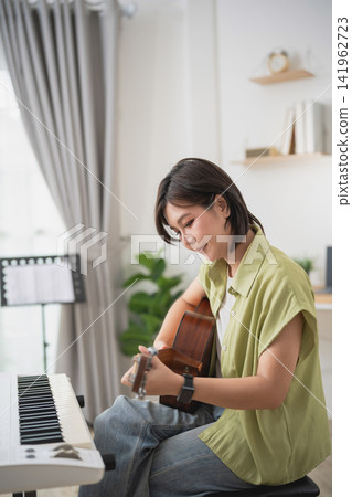 Woman playing acoustic guitar in bright modern living room with keyboard and bookshelf, relaxing and enjoying music Woman playing acoustic guitar in bright modern living room with keyboard and bookshelf, relaxing and enjoying music 141962723