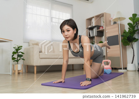 Young woman practicing yoga or stretching in living room on purple mat, wearing sportswear, with natural light coming through large windows 141962739