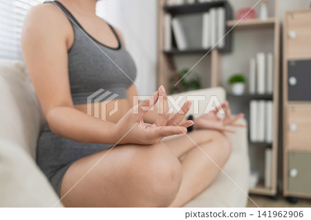 Close-up of a woman practicing meditation in a cozy living room, focusing on her hands in a mudra gesture while seated on a sofa Close-up of a woman practicing meditation in a cozy living room, focusing on her hands in a mudra gesture while seated on a sofa 141962906