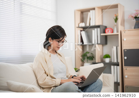 Woman Using Laptop in Cozy Home Office Setting with Bookshelf and Natural Light Woman Using Laptop in Cozy Home Office Setting with Bookshelf and Natural Light 141962910