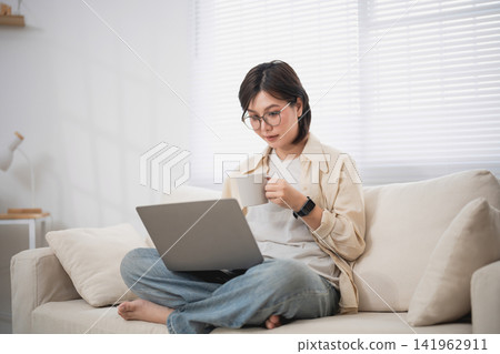 Woman Relaxing on Couch with Laptop and Drinking Coffee in Bright Living Room Woman Relaxing on Couch with Laptop and Drinking Coffee in Bright Living Room 141962911