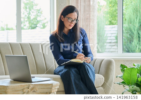 Attentive businesswoman taking notes in notebook while looking at laptop screen at home office Attentive businesswoman taking notes in notebook while looking at laptop screen at home office 141963158