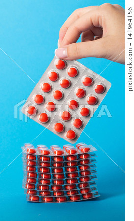 Human hand holding red pills blister pack above a high stack of medical drugs on solid blue background for pharmacy and healthcare concepts and medicine industry, vertical photo Human hand holding red pills blister pack above a high stack of medical drugs on solid blue background for pharmacy and healthcare concepts and medicine industry, vertical photo 141964156