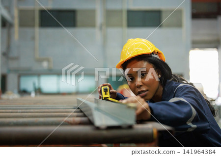 Beautiful African American female industrial worker in blue reflective uniform and hard hat working in factory using measuring tape. Beautiful African American female industrial worker in blue reflective uniform and hard hat working in factory using measuring tape. 141964384