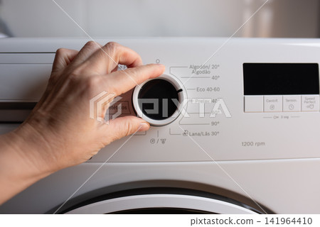 A hand adjusts the circular program selection knob on a white washing machine with Russian text. A hand adjusts the circular program selection knob on a white washing machine with Russian text. 141964410