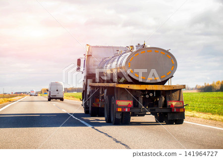 Large tanker truck travels asphalt highway under cloudy sky, hauling cylindrical container, commercial vehicle transport of liquids, freight delivery on open road, bright sun flare visible. Large tanker truck travels asphalt highway under cloudy sky, hauling cylindrical container, commercial vehicle transport of liquids, freight delivery on open road, bright sun flare visible. 141964727