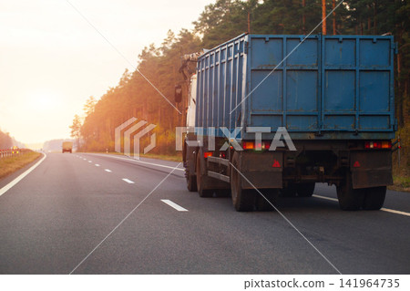 Rear view of large blue cargo truck traveling on asphalt highway, showing vehicle taillights with pine forest and autumn trees lining road under warm light of sunset or sunrise Rear view of large blue cargo truck traveling on asphalt highway, showing vehicle taillights with pine forest and autumn trees lining road under warm light of sunset or sunrise 141964735