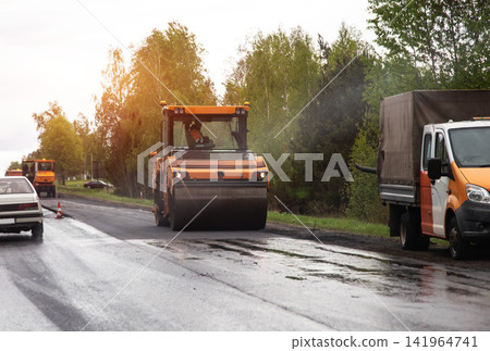 Road roller compacting fresh asphalt layer during construction work on suburban highway. Cargo truck parked at roadside, cars driving past. Copy space for text Road roller compacting fresh asphalt layer during construction work on suburban highway. Cargo truck parked at roadside, cars driving past. Copy space for text 141964741