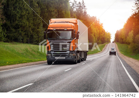 Large orange gray semi truck with white trailer travels country highway through green forest at golden hour sunset sunrise light Large orange gray semi truck with white trailer travels country highway through green forest at golden hour sunset sunrise light 141964742