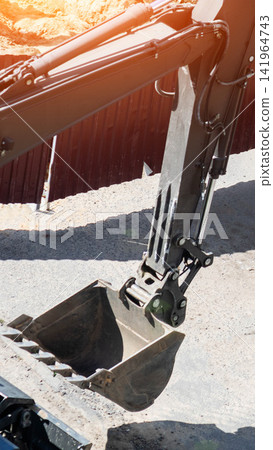 Excavator arm and bucket at construction site on sunny day, heavy machinery preparing ground for building development or infrastructure project with bright sun flare. Vertical photo Excavator arm and bucket at construction site on sunny day, heavy machinery preparing ground for building development or infrastructure project with bright sun flare. Vertical photo 141964743