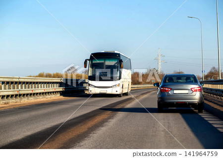 White tour bus traveling on bridge roadway towards camera perspective alongside grey car from rear. Clear blue sky and bare trees frame background scene. White tour bus traveling on bridge roadway towards camera perspective alongside grey car from rear. Clear blue sky and bare trees frame background scene. 141964759