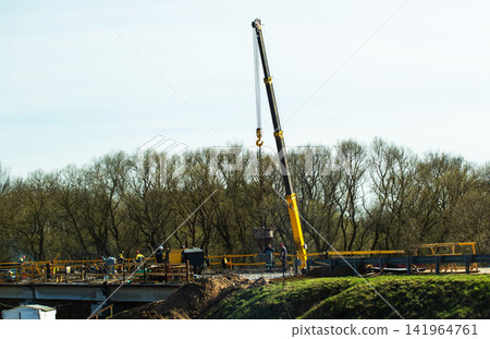 Construction of a bridge with a large yellow crane in operation, workers on structure, and bare trees in background under clear sky Construction of a bridge with a large yellow crane in operation, workers on structure, and bare trees in background under clear sky 141964761