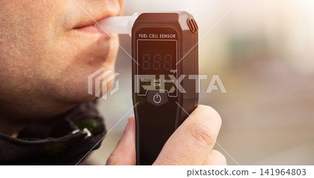 Man Blowing Into Breathalyzer Mouthpiece Outdoors. Closeup Of Male Driver Performing Alcohol Test. Man Blowing Into Breathalyzer Mouthpiece Outdoors. Closeup Of Male Driver Performing Alcohol Test. 141964803