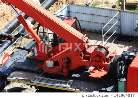 Red truck crane with extended boom and hydraulic cylinders performs heavy lifting operations at construction site trench with large black pipes and dirt under daylight Red truck crane with extended boom and hydraulic cylinders performs heavy lifting operations at construction site trench with large black pipes and dirt under daylight 141964817